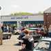 Outside of the cafe with cars parked up and people stood around talking.