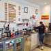 Inside the café the kitchen area behind the till with a coffee machine, fridges and the menu on the back wall.