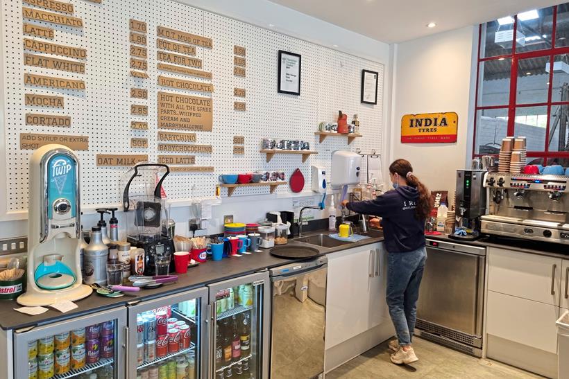 Inside the café the kitchen area behind the till with a coffee machine, fridges and the menu on the back wall.
